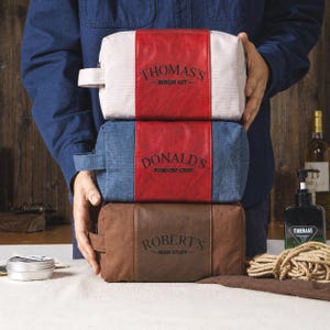 May include: Three rectangular toiletry bags stacked vertically. The top bag is cream with a red centre stripe labelled "THOMAS'S WASH KIT". The middle bag is blue with a red centre stripe labelled "DONALD'S RANDOM CRAP". The bottom bag is brown with a brown centre stripe labelled "ROBERT'S MAN STUFF".