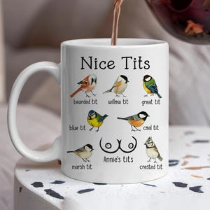May include: White ceramic mug with the text "Nice Tits" and illustrations of various bird species, including bearded tit, willow tit, and great tit. The mug also features the text "Annie's tits".