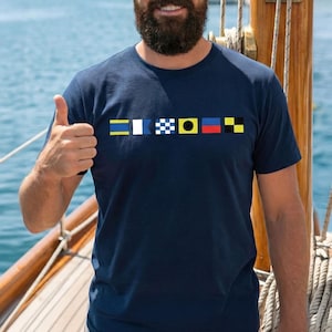 Peut inclure: T-shirt bleu marine orné d'une série de pavillons de signalisation nautiques colorés. Les drapeaux sont jaunes, blancs, bleus, noirs, rouges et jaunes. Le t-shirt est fait d'une matière douce.