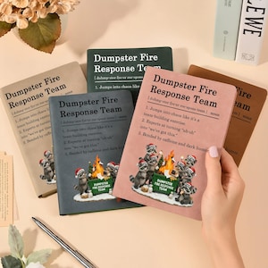 May include: A collection of notebooks with the text "Dumpster Fire Response Team" in various colors. Each notebook features a definition and a graphic of raccoons around a dumpster fire. The notebooks are a humorous office or gift item.
