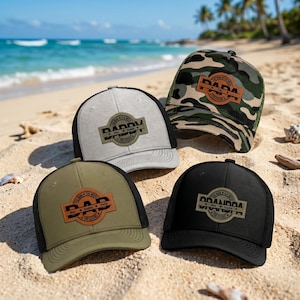 Può includere: Quattro cappellini da camionista con schienali in rete e visiere curve sono esposti su una spiaggia sabbiosa. I cappelli sono in vari colori: grigio, verde oliva, nero e mimetico. Ogni cappello presenta una toppa in pelle con le parole "DAD" o "GRANDPA".