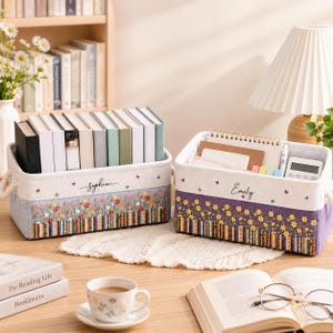 May include: Two rectangular fabric storage bins with rope handles, one light blue and one purple, featuring embroidered floral patterns and a row of book spines at the base. The bins are personalized with the names Sophia and Emily in black script.