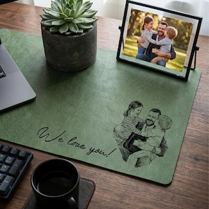 Könnte beinhalten: Grüne Schreibtischunterlage mit dem Text "We love you!" und einer Skizze einer umarmenden Familie. Auf dem Schreibtisch befinden sich ein gerahmtes Foto einer Familie, eine Sukkulente in einem Betontopf, ein Laptop, eine Tastatur und eine schwarze Kaffeetasse.