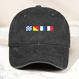 Op de afbeelding: Een donkergrijze baseballcap met een gebogen klep. De cap heeft een reeks kleurrijke nautische vlaggen. De vlaggen omvatten blauw-witte geblokte, geel-rode, wit-blauwe en rood-witte ontwerpen.