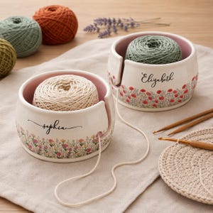 May include: Two round ceramic yarn bowls with a pink interior and a white exterior featuring a floral meadow pattern. One bowl is personalised with the name Sophia and the other with Elizabeth. Each bowl has a side cutout for yarn.