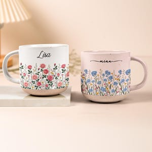 Könnte beinhalten: Zwei Keramikbecher mit gesprenkelten cremeweißen Basen und Henkeln. Der linke Becher zeigt rosa Rosen und den Namen Lisa in schwarzer Schreibschrift. Der rechte Becher zeigt blaue Wildblumen und den Namen mina in schwarzer Schreibschrift.