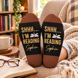 May include: A pair of black socks with brown heels and toes, featuring the yellow text SHHH... I'M READING, an illustration of an open book, and the name Sophia in cursive script. The socks are displayed side by side against a soft background.
