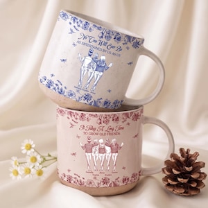 May include: Two ceramic mugs with speckled bases. The top mug is white with blue floral patterns and text reading No One Will Ever Be AS ENTERTAINED BY US AS US with names Isabella, Sophia, Anna. The bottom mug is pink with red floral patterns.