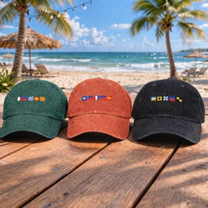 May include: Three corduroy baseball caps in green, rust, and black, each embroidered with colorful nautical signal flags. The caps are displayed on a wooden surface with a beach and ocean backdrop.
