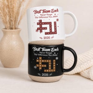 May include: Two ceramic mugs, one white and one black, each featuring a crossword-style design with the names Daniel, Sarah, Jack, Lind, and Tracy. The text reads Best Team Ever, Never Forget The Difference You Make, and the year 2026.