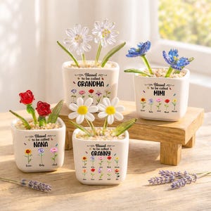 May include: Four small, square ceramic pots with decorative flowers. Each pot is white and features the phrase "Blessed to be called" followed by "GRANDMA", "MIMI", "NANA", and "GRANNY". The pots contain colorful, artificial flowers.