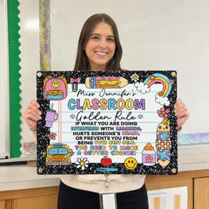 May include: A rectangular classroom sign with a black and white speckled border. The center features lined paper with the text: Miss Jennifer's CLASSROOM Golden Rule. IF WHAT YOU ARE DOING INTERFERES WITH LEARNING, HURTS SOMEONE'S HEART, OR PREVENTS YOU FROM BEING YOUR BEST SELF, YOU NEED TO MAKE A BETTER CHOICE! Decorated with colorful illustrations of a backpack, pencil, rainbow, school bus, apple, and smiley face.