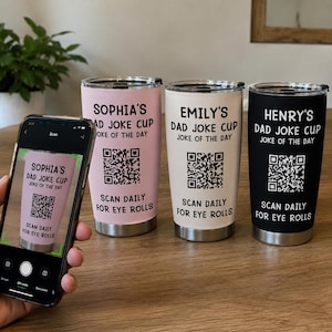 May include: Three stainless steel tumblers in light pink, cream, and black, each featuring a black QR code and the printed text: [Name]'s DAD JOKE CUP, JOKE OF THE DAY, SCAN DAILY FOR EYE ROLLS. A smartphone displays the pink tumbler on its screen.