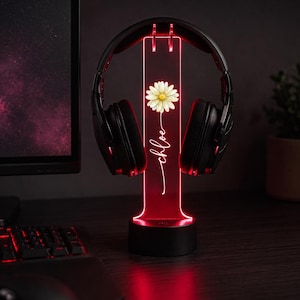 May include: A black headset resting on a vertical, rectangular, red-lit acrylic stand. The stand features a white daisy graphic above the name "chloe" written in a cursive script. The base is a solid black cylinder that illuminates the acrylic.