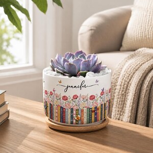Könnte beinhalten: Ein weißer, zylindrischer Keramikübertopf mit einer bunten Illustration einer Bücherreihe, die mit Blumen und Schmetterlingen verziert ist. Der Name Jennifer ist in schwarzer Schreibschrift aufgedruckt. Eine violette Sukkulente steht auf einem Holzsockel.