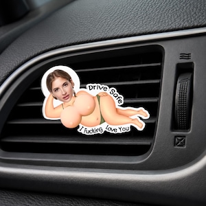 Peut inclure: Un désodorisant de voiture avec une illustration de dessin animé représentant une personne allongée en sous-vêtements verts avec un visage réaliste. Le texte Drive Safe et I Fucking Love You est imprimé en noir autour de la figure.