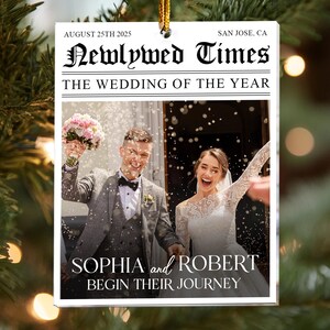 May include: A white rectangular ornament with a gold string for hanging. The ornament features a wedding photo of a smiling couple. Text on the ornament reads "Newlywed Times" and "The Wedding of the Year." The names "Sophia and Robert" are also displayed.
