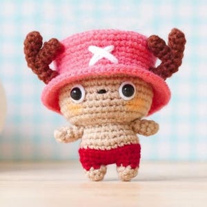 Puede incluir: Muñeco de ganchillo de un personaje de dibujos animados, con un gorro rosa con una cruz blanca, cuernos marrones y pantalones cortos rojos. El muñeco tiene grandes ojos negros y un cuerpo beige.