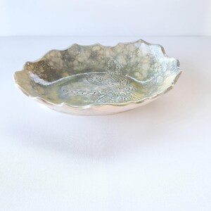 May include: An oval ceramic bowl with a scalloped edge. The bowl has a dragonfly design in the centre and is mainly green and beige. This decorative bowl is suitable for serving or display, and is a unique piece.