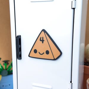 May include: A smiling, orange, triangular magnet with the number 4 and a black outline. The magnet is affixed to a white metal locker door. The magnet has two black dots for eyes and a curved black line for a mouth.