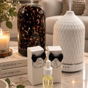 May include: Two essential oil diffusers, one with a dark tortoiseshell pattern and one with a white textured ceramic finish, emitting mist. Two white boxes with black bow ties labelled Clean Cotton and Musk & Tonka, and a glass dropper bottle.