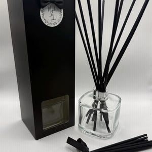 May include: A reed diffuser set featuring a clear square glass bottle with a silver collar, filled with black reeds. Beside it stands a tall black rectangular box with a circular label reading Myrrh & Tonka and a small black bow. Extra black reeds are tied with a black ribbon.