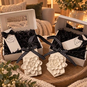 May include: Two white gift boxes filled with black shredded paper, each containing a white rectangular tag topped with a white sculpted flower and a black and white polka dot ribbon. One box includes a label reading Black Raspberry & Peppercorn.