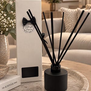 May include: A matte black cylindrical reed diffuser bottle with six black reeds, accompanied by a tall white rectangular box featuring a black bow and a circular label that reads Black Raspberry & Peppercorn in a decorative script font.