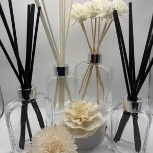 May include: Four clear glass rectangular diffuser bottles with silver collars, each containing either black or white reed sticks. Two bottles feature white decorative flower-shaped diffusers on top, while two others have bundles of straight reed sticks.