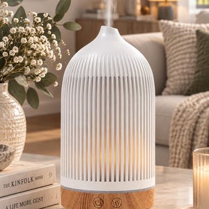 May include: A white, ribbed, dome-shaped essential oil diffuser with a light wood-grain base. The device features two circular control buttons on the front and emits a steady stream of mist from the top, indicating it is currently in operation.