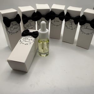 May include: A collection of six white rectangular boxes, each adorned with a black fabric bow and a circular label featuring floral details and text including Clean Cotton, Luna, and Cashmeran. A clear glass dropper bottle with yellow liquid sits in front.