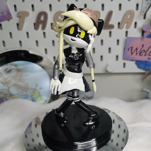 May include: A figurine of a character with a black and white outfit, yellow hair, and black accents. The figure is standing on a black circular base, which is on a mirrored surface. The background includes the word "NAZLA" in colorful letters.