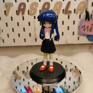 Peut inclure: Figurine d'un personnage aux cheveux bleus, avec un nœud rose et une jupe noire, debout sur une plateforme miroir. La figurine porte des chaussures rouges et des chaussettes blanches. Le fond affiche le texte "NAZLA TASARLA".