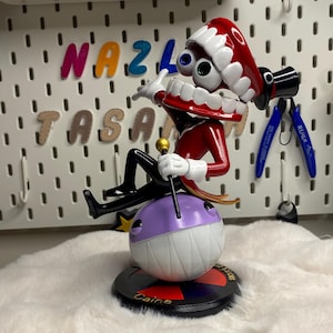 May include: A figurine of a character with a red and white hat, large teeth, and multiple eyes. The figure is perched on a purple and white ball, with a black base that reads "Caine". The background includes the text "NAZLI TASA".
