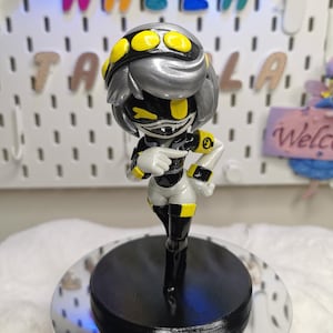 May include: A figurine of a character with a silver head, black face, and yellow accents. The figure is wearing a black and white outfit with yellow details. It stands on a black circular base atop a mirrored surface. The name "NAZLA" is displayed in colorful letters in the background.
