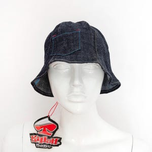 May include: Dark blue denim bucket hat with a small pocket and red and blue stitching details. The hat is displayed on a white mannequin head. A tag with the text "Killah Babe" hangs from the hat.