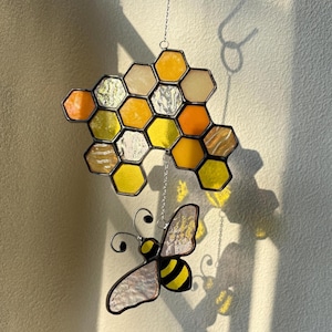 May include: Stained glass honeycomb and bee suncatcher. The honeycomb is made of yellow, orange, and clear glass. The bee has yellow and black stripes with iridescent wings. The suncatcher hangs from a chain.