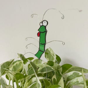 May include: A whimsical, green and black stained glass insect with wire antennae and legs, perched atop a lush green and white plant. The insect has a red tongue sticking out, adding a playful touch to the garden decor.