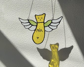May include: A yellow stained glass angel with white and green wings, hanging from a chain. The angel has the name "MIA" written on it. The angel's shadow is visible on the white wall.