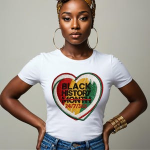 Pode incluir: Camiseta branca com um design bordado em forma de coração. O design inclui o texto "BLACK HISTORY MONTH 24/7/365" em preto, vermelho, amarelo e verde. A camiseta é de gola redonda.