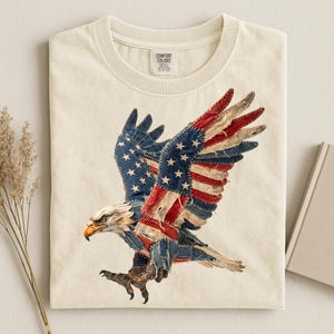 May include: Cream-colored t-shirt featuring a detailed graphic of an American bald eagle with wings spread. The eagle's wings are designed with the American flag's stars and stripes. The shirt has a crew neck and a tag that says "Comfort Colors".