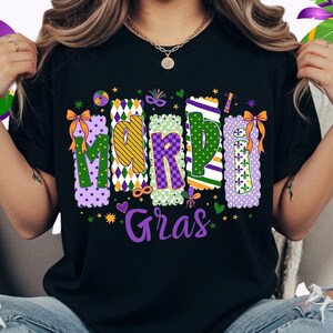May include: Black t-shirt with a colorful "Mardi Gras" design. The text is in a decorative font with purple, green, and gold accents. The design includes stars, bows, and decorative elements, perfect for celebrating the festive season.