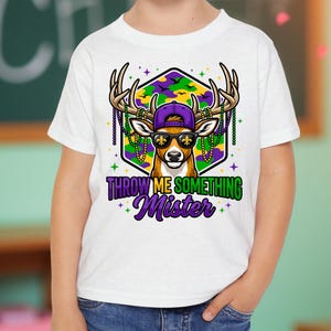 May include: White t-shirt with a deer graphic wearing sunglasses and a purple cap. The deer is decorated with beads and antlers, set against a geometric background. The text "THROW ME SOMETHING Mister" is displayed below the deer.