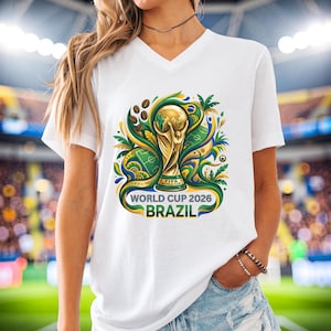 May include: White V-neck t-shirt with a vibrant graphic of the FIFA World Cup trophy, soccer ball, and green and yellow details. The text "WORLD CUP 2026 BRAZIL" is printed below the trophy.