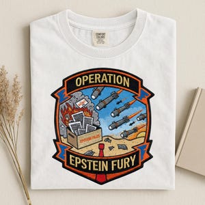 May include: White t-shirt with a graphic patch reading "OPERATION EPSTEIN FURY". The patch shows a burning building, missiles, and a box labelled "EPSTEIN FILES". The shirt has a crew neck and a Comfort Colors label.