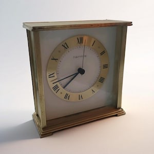 Könnte beinhalten: Eine Vintage-Uhr in Goldtönen mit weißem Zifferblatt und schwarzen Zeigern. Das Zifferblatt hat goldene römische Ziffern und den Markennamen "hermie". Die Uhr ist rechteckig mit einem goldenen Rahmen und einer Glasfront.