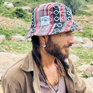 Könnte beinhalten: Ein bunter Fischerhut mit einem geometrischen Muster in Rot, Schwarz und Weiß. Der Hut hat ein Etikett mit der Aufschrift "Himalayan Hemp". Die Person trägt auch eine braune Cordjacke.