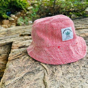 Può includere: Cappello a secchiello rosso e bianco con motivo a spina di pesce. Ha una piccola etichetta con la scritta "Himalayan Hemp THC Free Made in India". Il cappello è realizzato in tessuto strutturato ed è appoggiato su una superficie rocciosa.