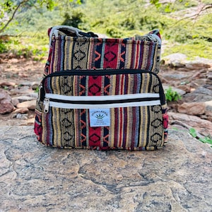 May include: A colorful, patterned shoulder bag with a front zippered pocket. The bag features a mix of red, black, yellow, and white stripes and geometric designs. A small tag reads "Himalayan Hemp". The bag is sitting on a rock.