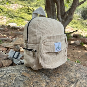 May include: A small, beige backpack with a herringbone pattern. The backpack has a front pocket, black zips, and a small handle. The bag is made of a woven material and sits on a rock. The backpack has a small tag with text and a logo.